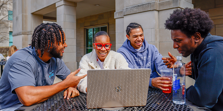 4 Black students sitting around a table outside gesturing toward a laptop.